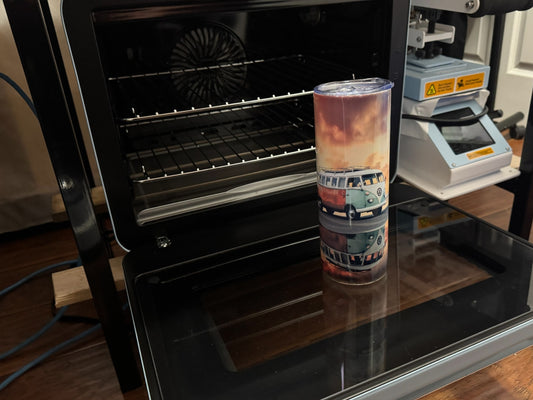 A sublimation tumbler with a vibrant design of a vintage van placed on top of a sublimation oven, with a heat press machine visible in the background.